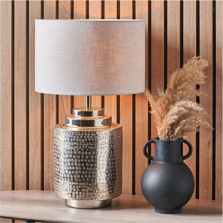 Zuri Silver Metal Table Lamp with a textured dotted pattern base and white linen cylinder shade, shown on a console table beside a black vase with dried pampas grass.