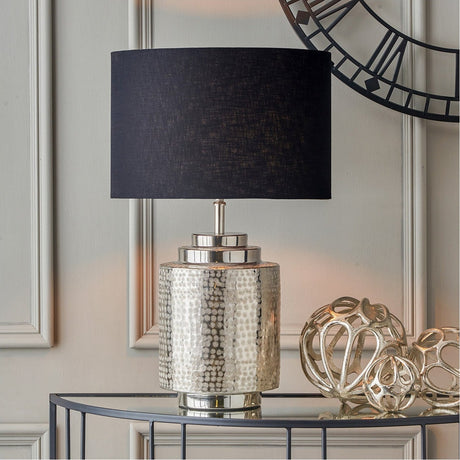 Zuri Shiny Silver Aluminium Pot Table Lamp with textured cylindrical base and black drum shade, styled on a glass console table beside decorative metal ornaments.
