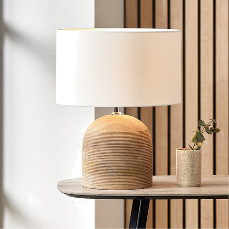 Nelu table lamp with a ridged natural wood dome base and white handloom cylinder shade, shown on a wooden side table beside a small potted plant.