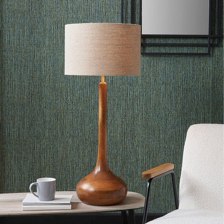 Toma Oiled Wood Tall Neck Table Lamp Base with a linen drum shade, styled on a wooden side table beside books and a mug in a living room with green textured wallpaper.