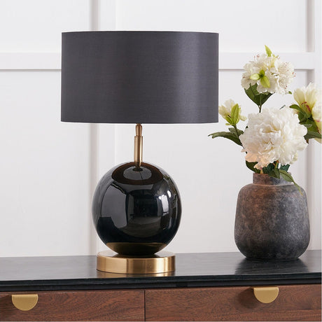 Sofia Black & Gold Enamel Table Lamp Base with round glossy black glass body, brass stem and base, paired with a grey drum shade, shown on a dark sideboard.