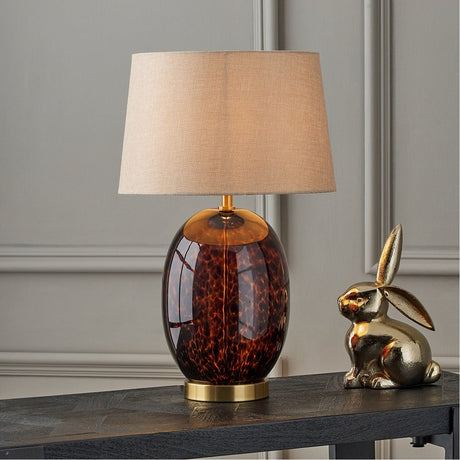 Lucien Tortoiseshell Brown Glass Tall Table Lamp with a beige linen drum shade and brass fittings, styled on a dark console table beside a gold rabbit figurine.