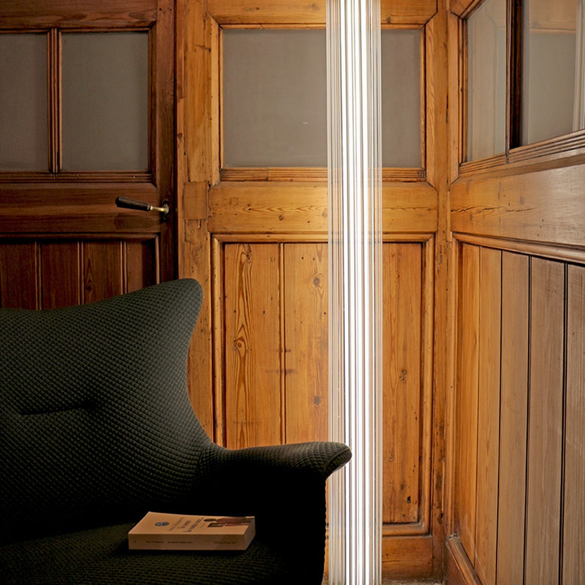 Dalvorne Glass Floor Lamp in satin bronze, a tall ribbed glass column lighting a wood-panelled room beside a dark upholstered armchair.