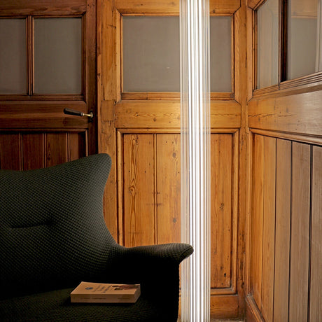 Dalvorne Glass Floor Lamp in satin bronze, a tall ribbed glass column lighting a wood-panelled room beside a dark upholstered armchair.
