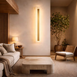 A minimalist living room in neutral tones features a beige sofa with cushions, a low square coffee table, a woven chair, a potted tree, and soft lighting from the Averra LED Large Rectangle Alabaster Wall Light in Soft White & Brass and a small table lamp.