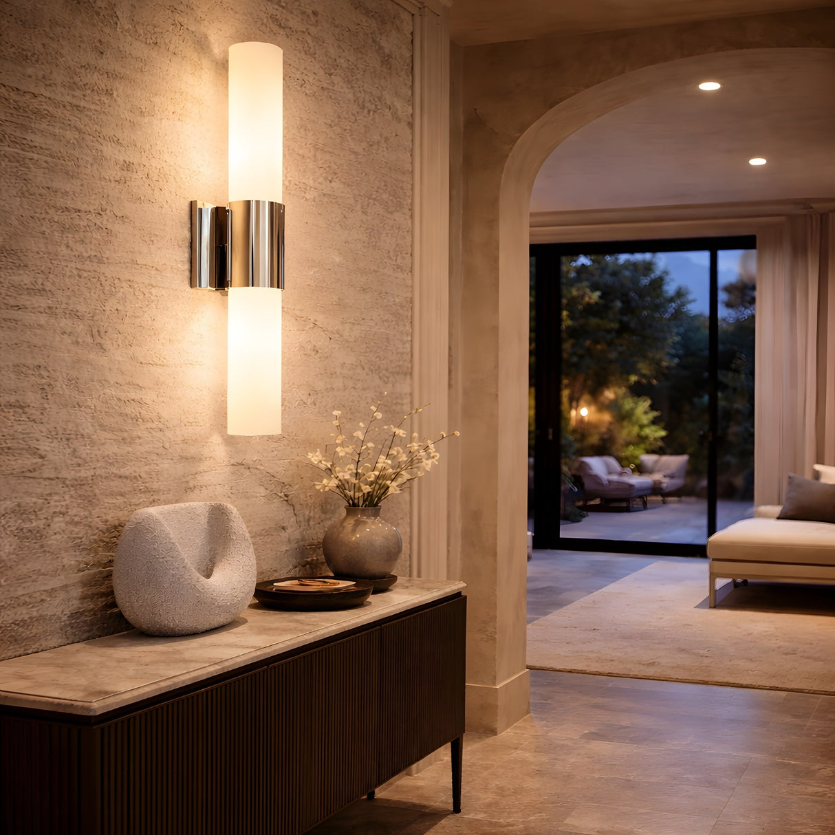A modern hallway features a textured wall, Fiora LED Alabaster Wall Light – Satin Nickel & Soft White, decorative vases, and a sculptural object on a console table; an archway leads to a living room with sofa and glass doors opening to the outdoor area at dusk.