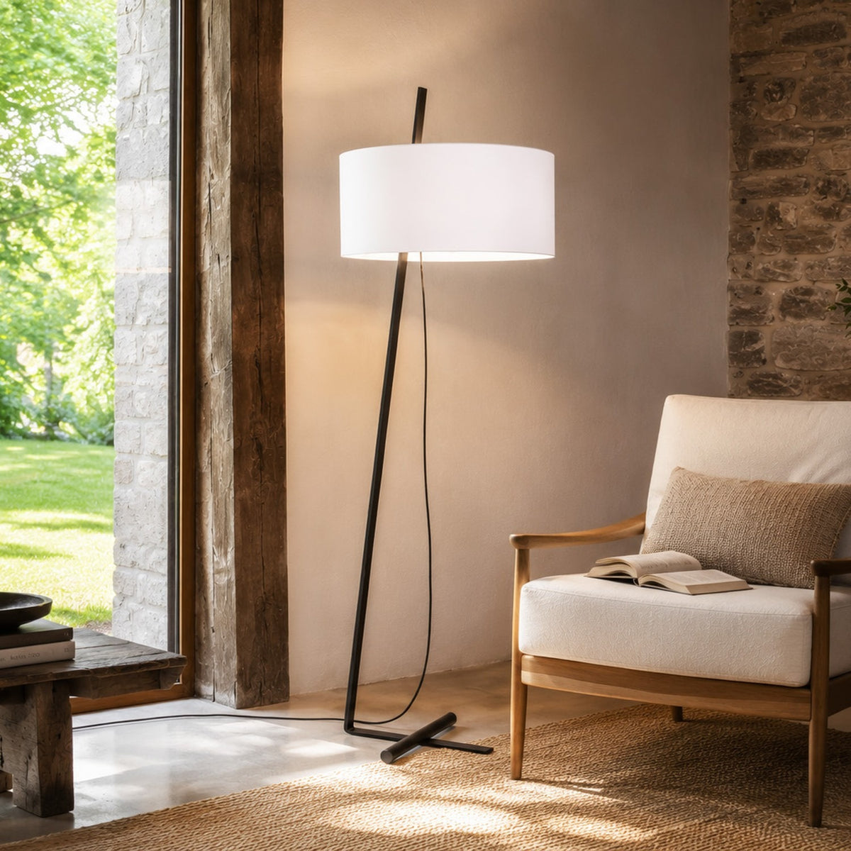 Gilvorne fabric floor lamp with white cotton drum shade on a slanted anthracite iron stem and cross-shaped base, shown beside an armchair in a living room.