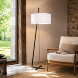 Gilvorne fabric floor lamp with white cotton drum shade on a slanted anthracite iron stem and cross-shaped base, shown beside an armchair in a living room.