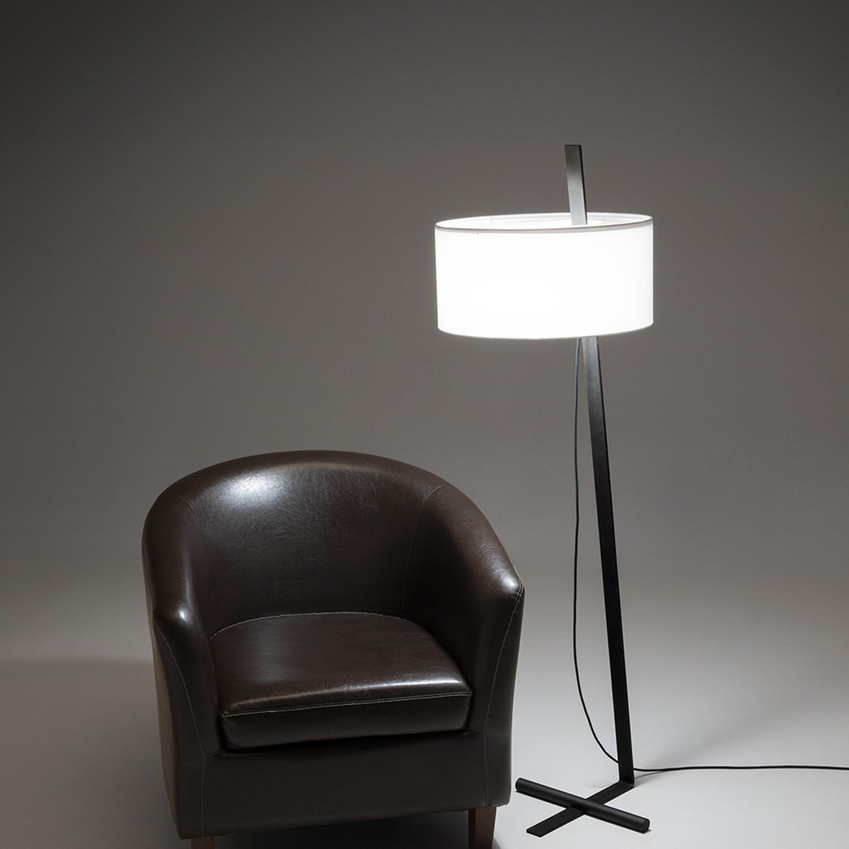 Gilvorne Fabric Floor Lamp in anthracite and white, featuring a slim angled iron stem, cross-shaped base, and broad white cotton drum shade, shown lit beside a brown leather armchair.