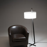 Gilvorne Fabric Floor Lamp in anthracite and white, featuring a slim angled iron stem, cross-shaped base, and broad white cotton drum shade, shown lit beside a brown leather armchair.