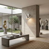 A modern, minimalist art gallery features abstract sculptures on pedestals, large windows overlooking a courtyard, neutral-toned decor bathed in natural light, and white Funk Wall Lights accenting the walls.