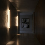 A dimly lit hallway features Averra LED Medium Rectangle Alabaster Wall Lights in Soft White & Brass on the left, a dark wall to the right, and an abstract black and white painting at the far end.