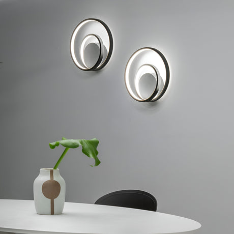 A minimalist room is elegantly designed with a white oval table showcasing a modern vase that holds a single green leaf. Mounted on the light gray wall are two Glistis Dimmable LED Wall Lights in black, each offering 28W of energy-efficient illumination and featuring metal accents. These lights contribute to an elegant and contemporary atmosphere.