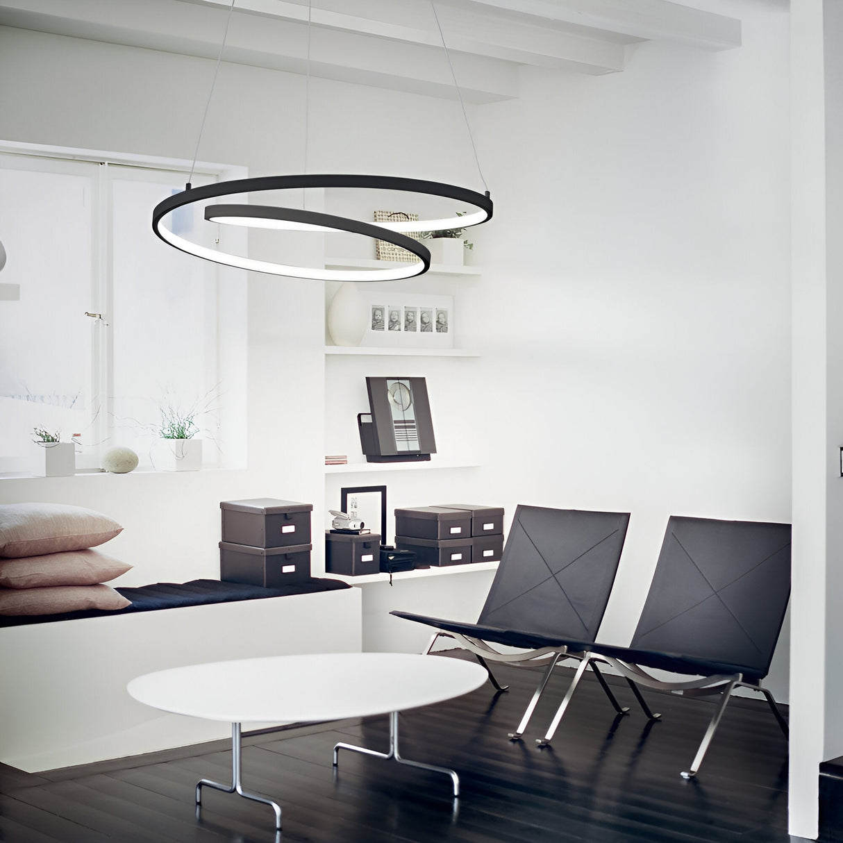 A minimalist room showcases a modern aesthetic with two black chairs and a white coffee table on dark wood flooring. The Glistis Dimmable LED Suspension Light 3000K, 55W - Black, 80cm hangs above, enhancing the ambiance. Shelves with decorative items and a window nook adorned with pillows complete the backdrop.