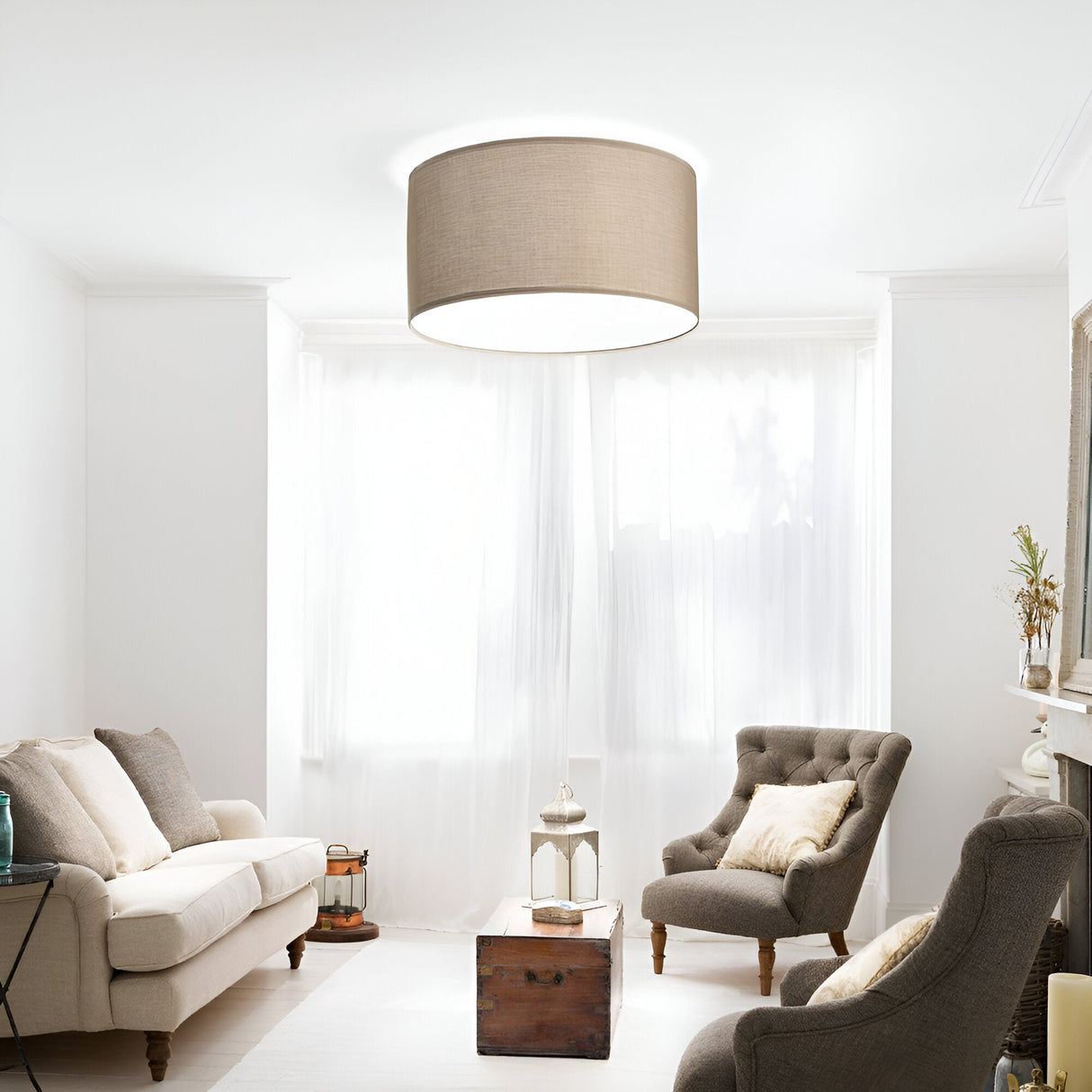 A cozy living room with a neutral color palette features a beige sofa and gray armchair, both adorned with cushions. Overhead, an Aethera Cylinder Lampshade in Dark Beige (70cm) lends elegance to the space. A small wooden coffee table sits in the center, while the mantel on the right displays timeless design decor.