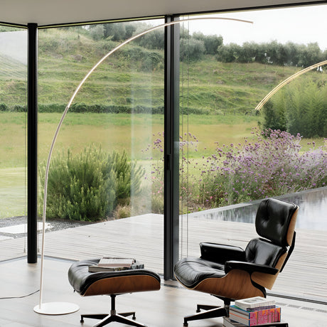 A modern living room features a black leather lounge chair and ottoman near a large glass window overlooking a lush green landscape with hills and purple flowers. The Next LED Floor Lamp 30W 2000K in white offers the perfect modern lighting solution, while books are neatly stacked on the ottoman, enhancing the contemporary look.