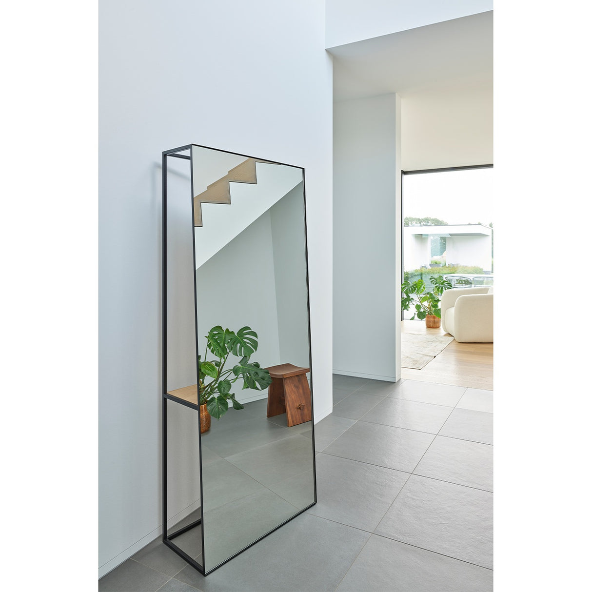 Chassis XL Oak Shelf Free Standing Mirror 170cm in Black, an open black metal frame cheval mirror with a solid oak shelf, shown leaning in a bright hallway with a plant and stool reflected.