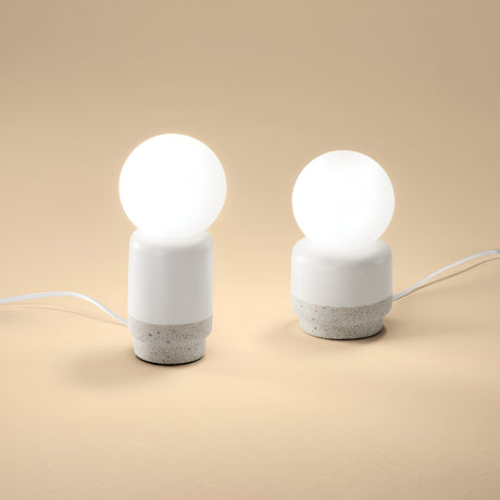Two Brillara Large Table Lamps in grey, featuring round white globes, stand on a beige surface. Each lamp displays a textured ceramic base that ingeniously mimics the appearance of cement. Offering ambient lighting, their cords are discreetly hidden from view as they illuminate the room with a gentle glow.