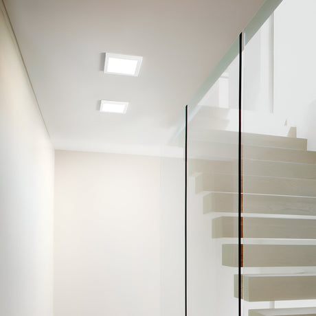 A modern interior showcases a glass staircase with wooden steps on the right, divided by a clear glass partition. The ceiling is brightened by two Lucella Recessed Square LED Downlights at 4000K, emphasizing the minimalist design and neutral color palette while focusing on energy efficiency.