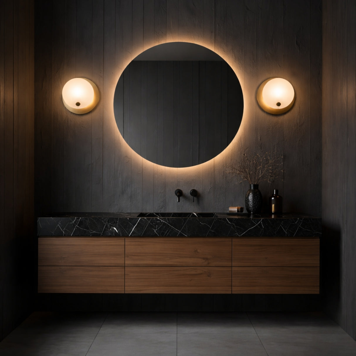 A modern bathroom with dark walls, a round backlit mirror, an Avelora LED Round Alabaster Wall Light in soft white and brass, a black marble countertop, wooden drawers, and minimalist decor like a vase with dried branches and bottles.