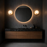 A modern bathroom with dark walls, a round backlit mirror, an Avelora LED Round Alabaster Wall Light in soft white and brass, a black marble countertop, wooden drawers, and minimalist decor like a vase with dried branches and bottles.