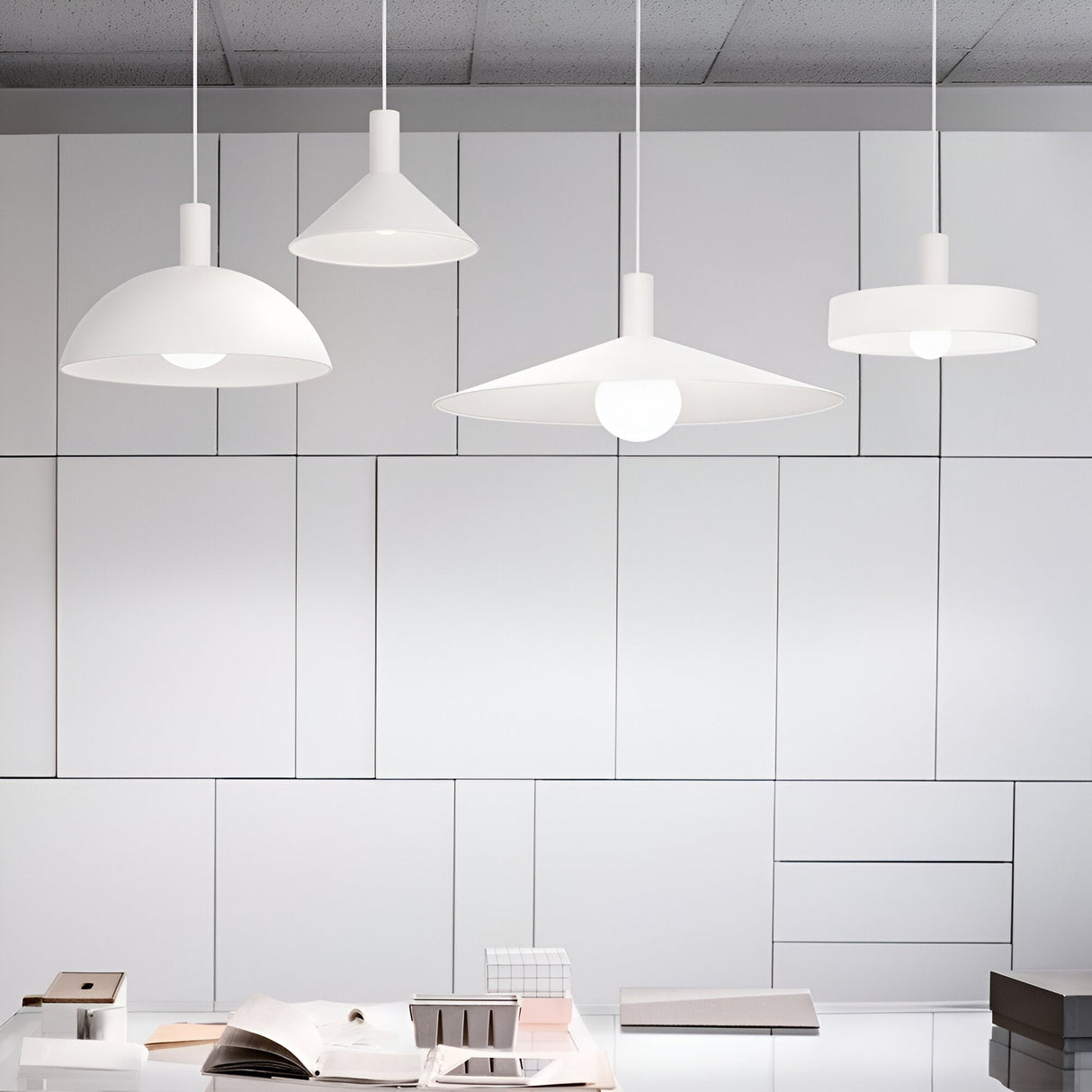 A modern, minimalistic office space is illuminated by four Arion Small Ceiling Lampshades in White, hanging gracefully from the ceiling to elevate the industrial-style interior. The background features a wall with geometric paneling, while the foreground displays a desk decorated with open books and essential office supplies.