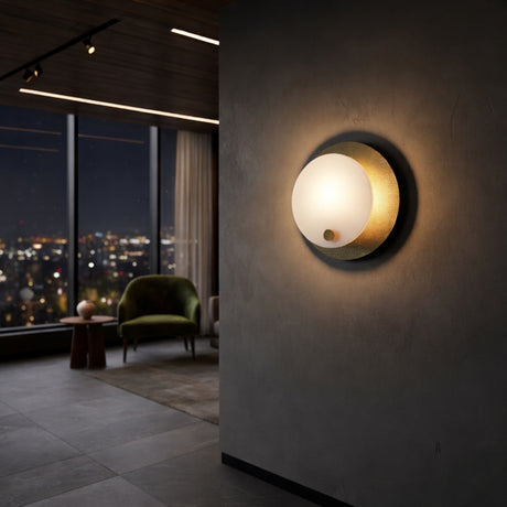 The Avelora LED Round Alabaster Wall Light in soft white & brass glows warmly in a stylish living room with large windows, city views, and a green armchair paired with a small table by the window.