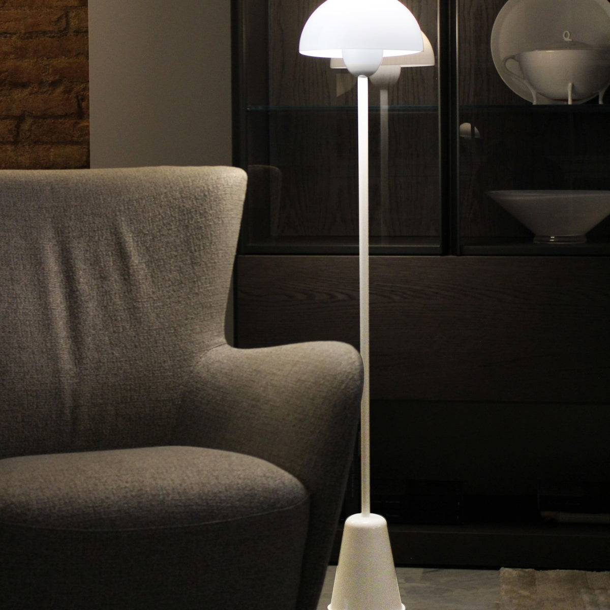 Star Floor Lamp in glossy white with a dome acrylic shade atop a slender iron stem and tapered conical base, lit beside a grey armchair in a living room setting.