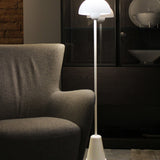 Star Floor Lamp in glossy white with a dome acrylic shade atop a slender iron stem and tapered conical base, lit beside a grey armchair in a living room setting.