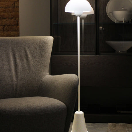 Star Floor Lamp in glossy white with a dome acrylic shade atop a slender iron stem and tapered conical base, lit beside a grey armchair in a living room setting.