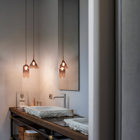 A modern bathroom features two stone sinks on a wooden countertop. Hanging overhead are three Empire-2 1 Light Glass Pendants in black, each compatible with E27 bulbs, providing illumination. A large mirror reflects the setup, while a small sculpture rests by the switch on the wall. Below, a wicker basket can be seen.