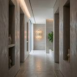 Magari 2 Light Crystal Glass Wall Light in gold illuminated at the end of a grey plaster hallway with built-in niches, vases, and a fiddle-leaf fig.
