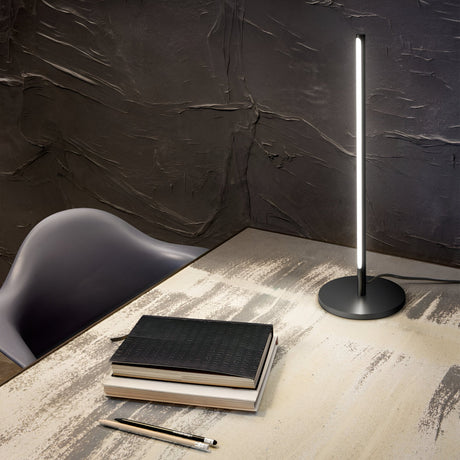 The Celestia LED Table Lamp in black casts a modern glow across the workspace, contrasting with the dark textured walls. On the wooden desk, a stack of books, a notebook, and a pencil are neatly arranged. A sleek black chair is partially visible on the left side.