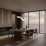 Niori Zareon curved alabaster LED chandelier in brushed brass suspended above a walnut dining table in a modern oceanfront room at dusk.