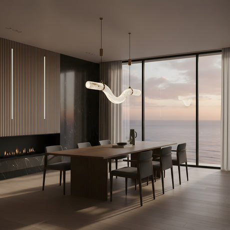 A modern dining room with a wooden table, eight chairs, the Zareon LED Medium Curved Alabaster Suspension Light in soft white, a wall-mounted fireplace, and floor-to-ceiling windows overlooking the ocean at sunset.
