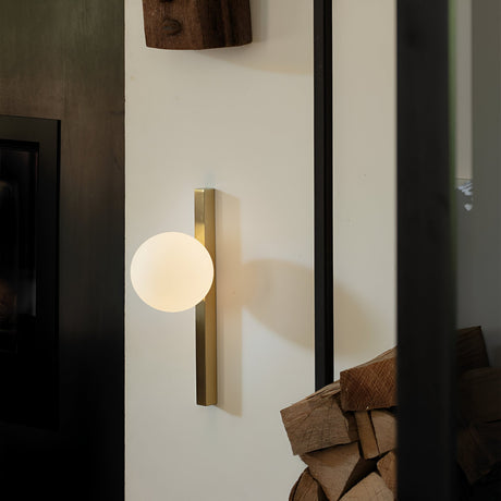 The Phostis Ceiling & Wall Light in gold is a modern classic, showcasing a round, glowing bulb and a vertical metallic base mounted on a white wall. Nearby, a stack of firewood complements the setting. The brushed brass finish casts intriguing shadows that add depth to the scene.