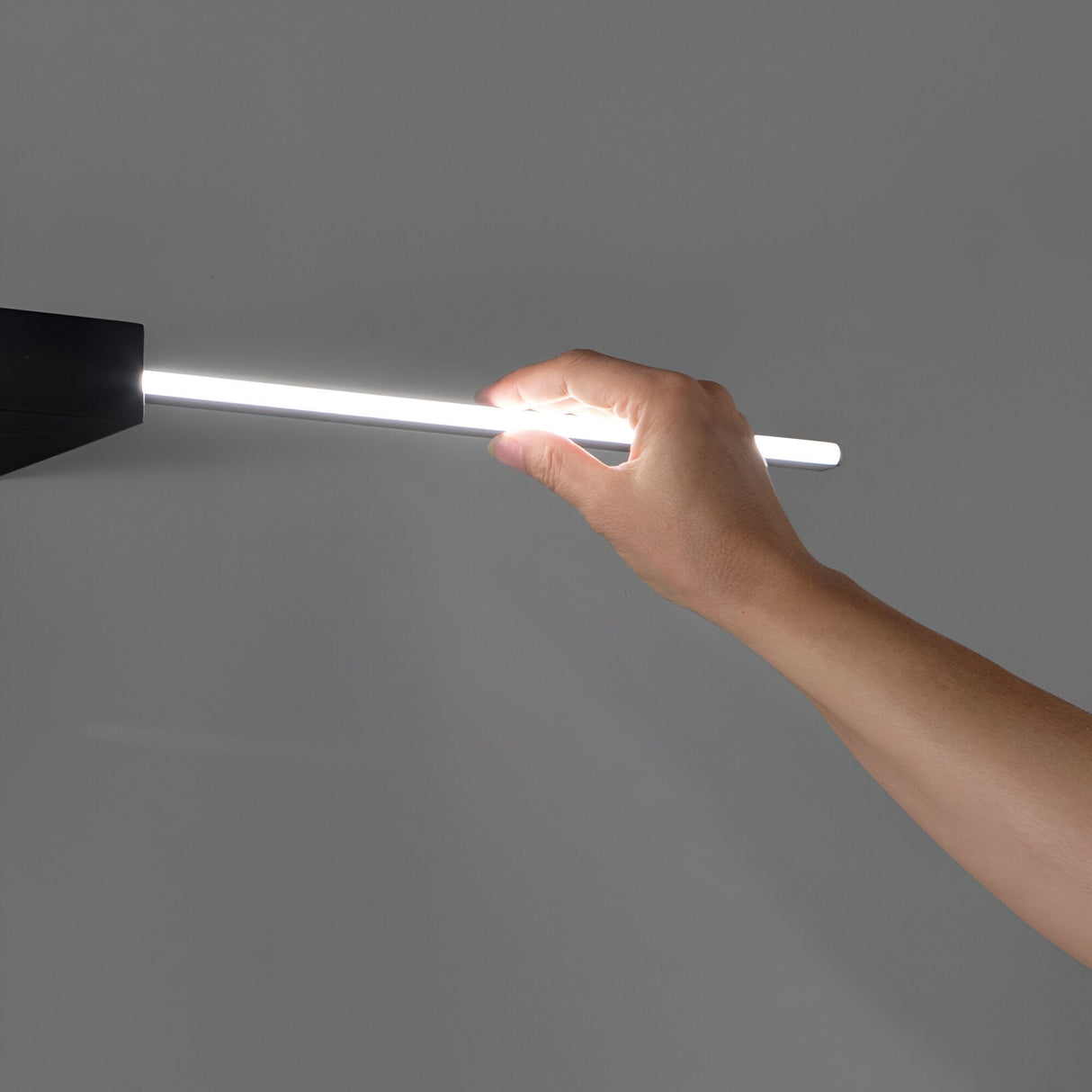 A hand holds the sleek Helion LED Picture Light 13.5W 3000K - Black, its glowing cylindrical form extending from a dark fixture against a plain gray background. This light emits a bright white glow, ideal for contemporary interiors, creating a subtle aura around the hand.