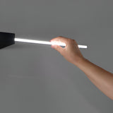 A hand holds the sleek Helion LED Picture Light 13.5W 3000K - Black, its glowing cylindrical form extending from a dark fixture against a plain gray background. This light emits a bright white glow, ideal for contemporary interiors, creating a subtle aura around the hand.