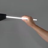 A hand is gently holding the Helion LED Picture Light 20.5W 3000K in black, featuring a matte finish and cylindrical design, attached to a wall fixture. The background is a plain gray wall, highlighting the light's brightness and elegant simplicity, making it ideal for illuminating refined artwork.