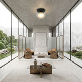 Minimalist modern room with glass walls, two tan chairs, a low table, and decorative screens. A Galaxy 1 Light Wall/Flush Ceiling Light - Black & White is centered above neutral furnishings. Sculpted greenery and rocks are seen outside.