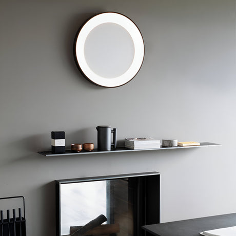 A modern interior showcases a sleek gray wall with a round, backlit mirror. The Solisca LED Ceiling & Wall Light 33W 3000K in Black, measuring 60cm, enhances the minimalist shelf below, which is adorned with books, a coffee pot, and small decor items. Beneath the shelf, a fireplace with a reflective surface complements the modern home lighting beautifully.