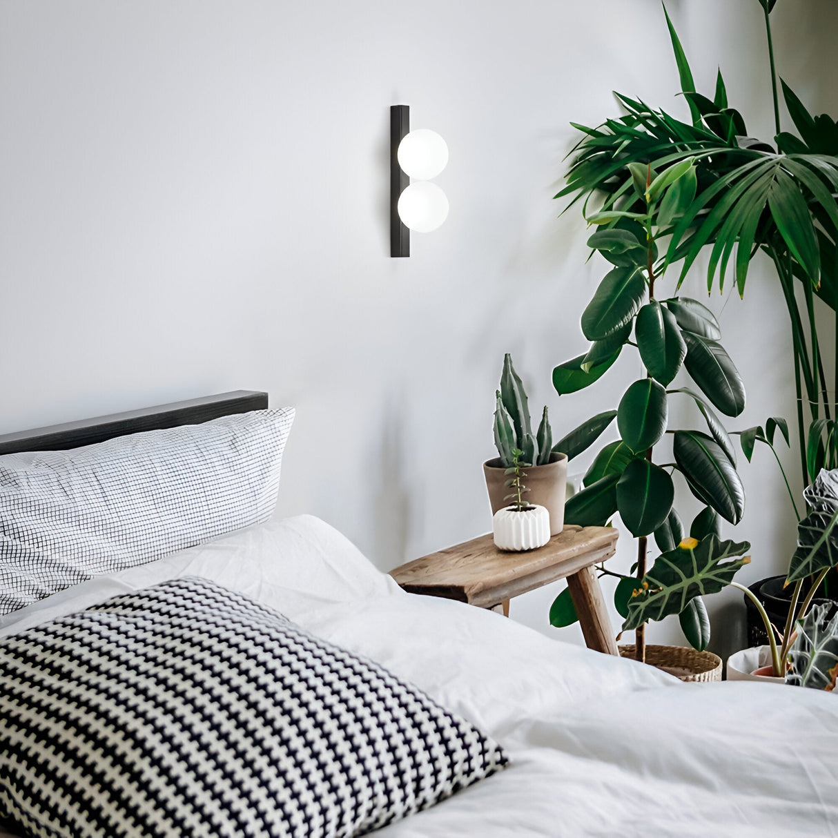 A cozy bedroom showcases a neatly made bed adorned with black and white checkered pillows, alongside a wooden side table displaying potted plants. The wall is accented by the modern Lyria 2 Light Ceiling & Wall Light 6W 3000K in black. A large leafy plant enhances the corner against the plain white wall, perfectly highlighting the contemporary lighting elements.