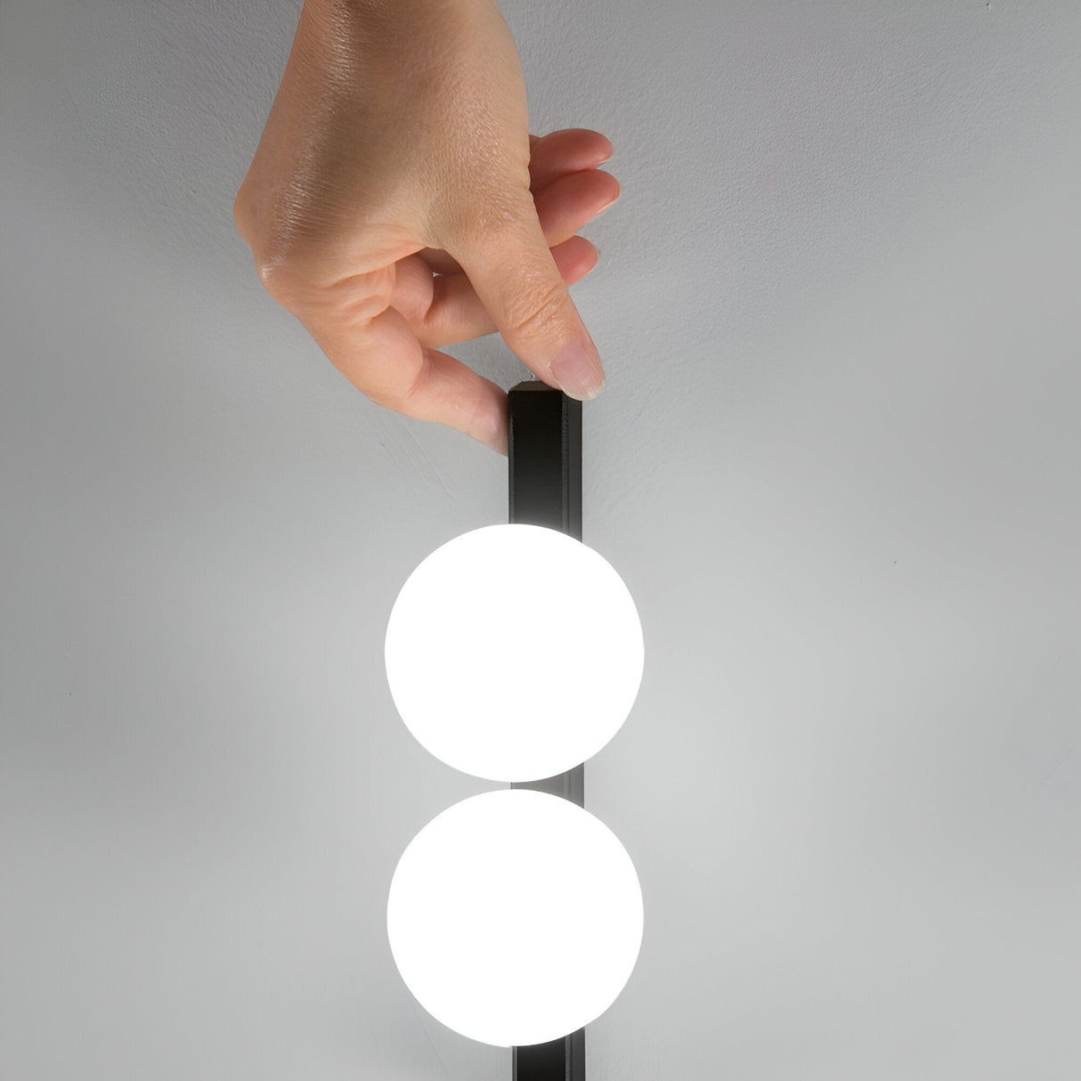 A hand adjusts the vertical Lyria 2 Light Ceiling & Wall Light 6W 3000K - Black, showcasing its two glowing circular bulbs against a plain white backdrop. Ideal for modern interiors, this fixture combines sleek design with energy-saving efficiency.