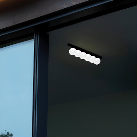 In a contemporary design, the Lyria 4 Light Wall Light 12W 3000K in Black presents a sleek arrangement of round bulbs, perfectly mounted on a dark wall beside a large glass door or window. The minimalist setting underscores the fixture's modern elegance against the reflective glass surface.