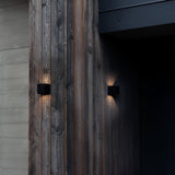 A contemporary wooden exterior wall with a pair of minimalist Flareon LED Outdoor 12W 3000K Wall Lights in black casts warm glows across its surface. The wood showcases natural grain patterns and various shades, providing a contrast to the dark entrance. These energy-efficient lights not only enhance the ambiance but also offer an eco-friendly lighting solution.