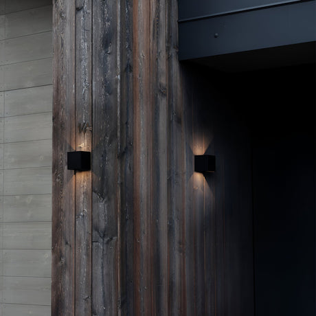 A contemporary wooden exterior wall with a pair of minimalist Flareon LED Outdoor 12W 3000K Wall Lights in black casts warm glows across its surface. The wood showcases natural grain patterns and various shades, providing a contrast to the dark entrance. These energy-efficient lights not only enhance the ambiance but also offer an eco-friendly lighting solution.