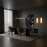 A modern dining room with a dark marble table, six black chairs, abstract wall art, an Avelora LED Small Capsule Alabaster Wall Light in soft white & brass, and a large window with sheer curtains. Decor is neutral-toned and minimalist.