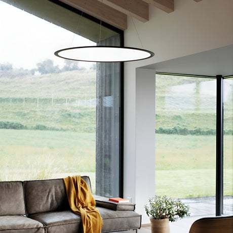 A modern living room showcases a gray sofa, a yellow throw, and the Luxra LED Suspension Light 61W 3000K in black, measuring 93cm, which casts ambient lighting. Expansive floor-to-ceiling windows reveal scenic green fields and hills outside. A potted plant rests near the window, enhancing the sleek modern design.