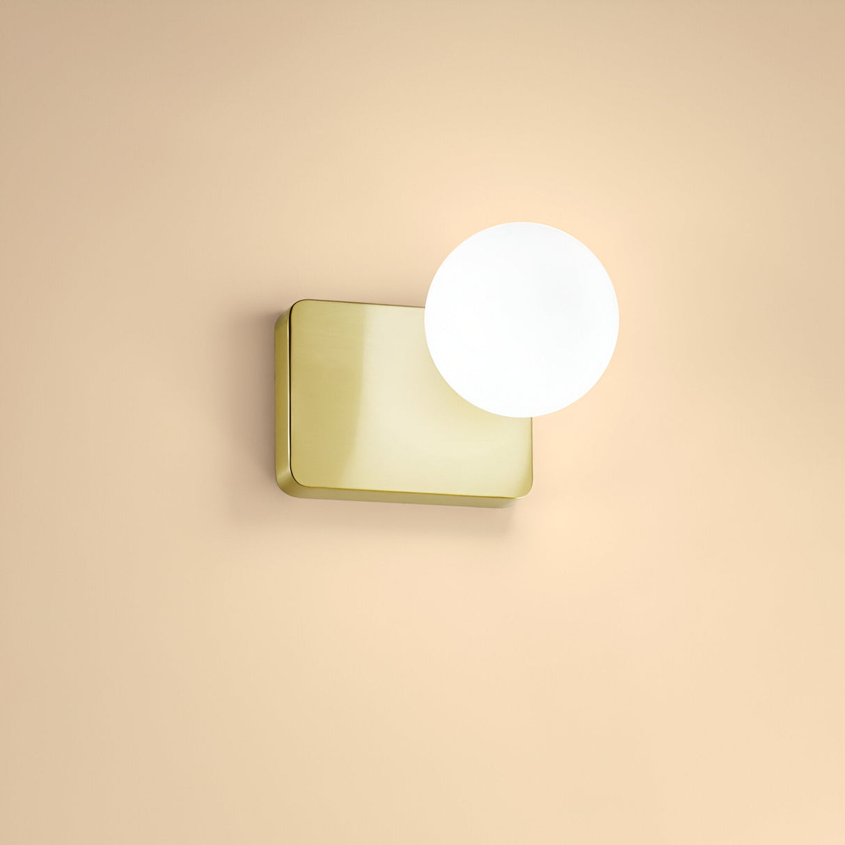 The Luxis 1 Light Wall Light in gold, featuring a minimalist design with a square gold-finish base and a glowing white spherical bulb, is elegantly mounted on a beige wall.