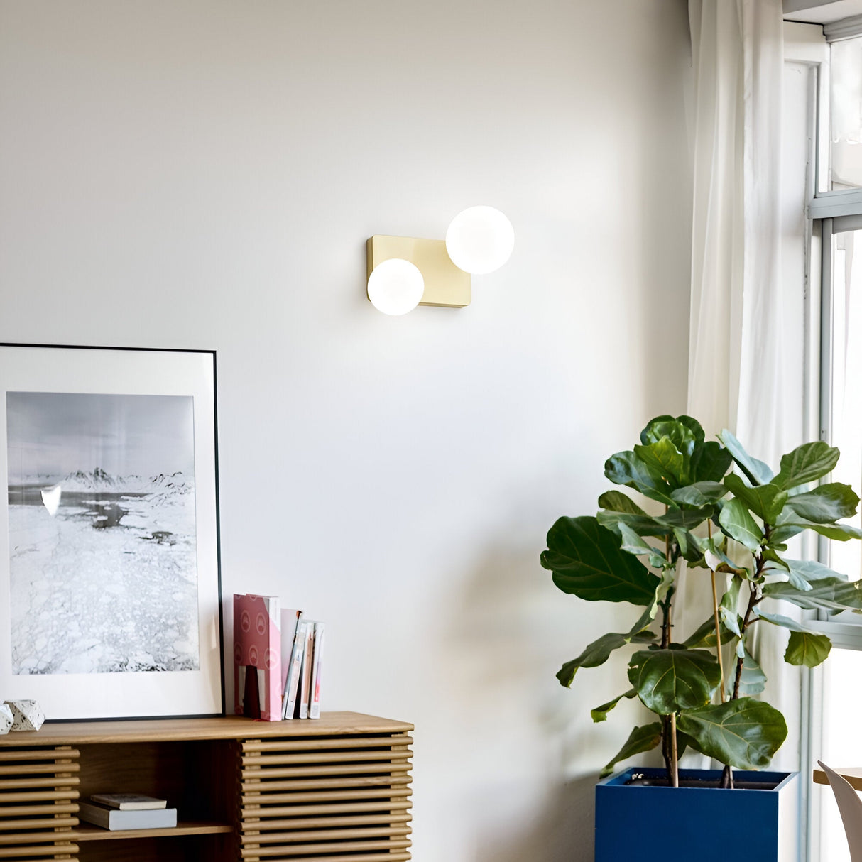 In a contemporary living room with a white wall, the Luxis 2 Light Wall Light - Gold provides an elegant glow. Beneath it is a wooden sideboard displaying a framed black-and-white photograph and several books. A large fiddle leaf fig plant stands beside a window adorned with white curtains, adding to the tranquil atmosphere.